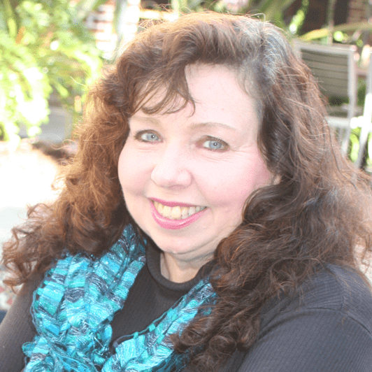 A smiling woman with curly brown hair and blue eyes wears a teal scarf. The background features greenery, suggesting a sunny outdoor setting.