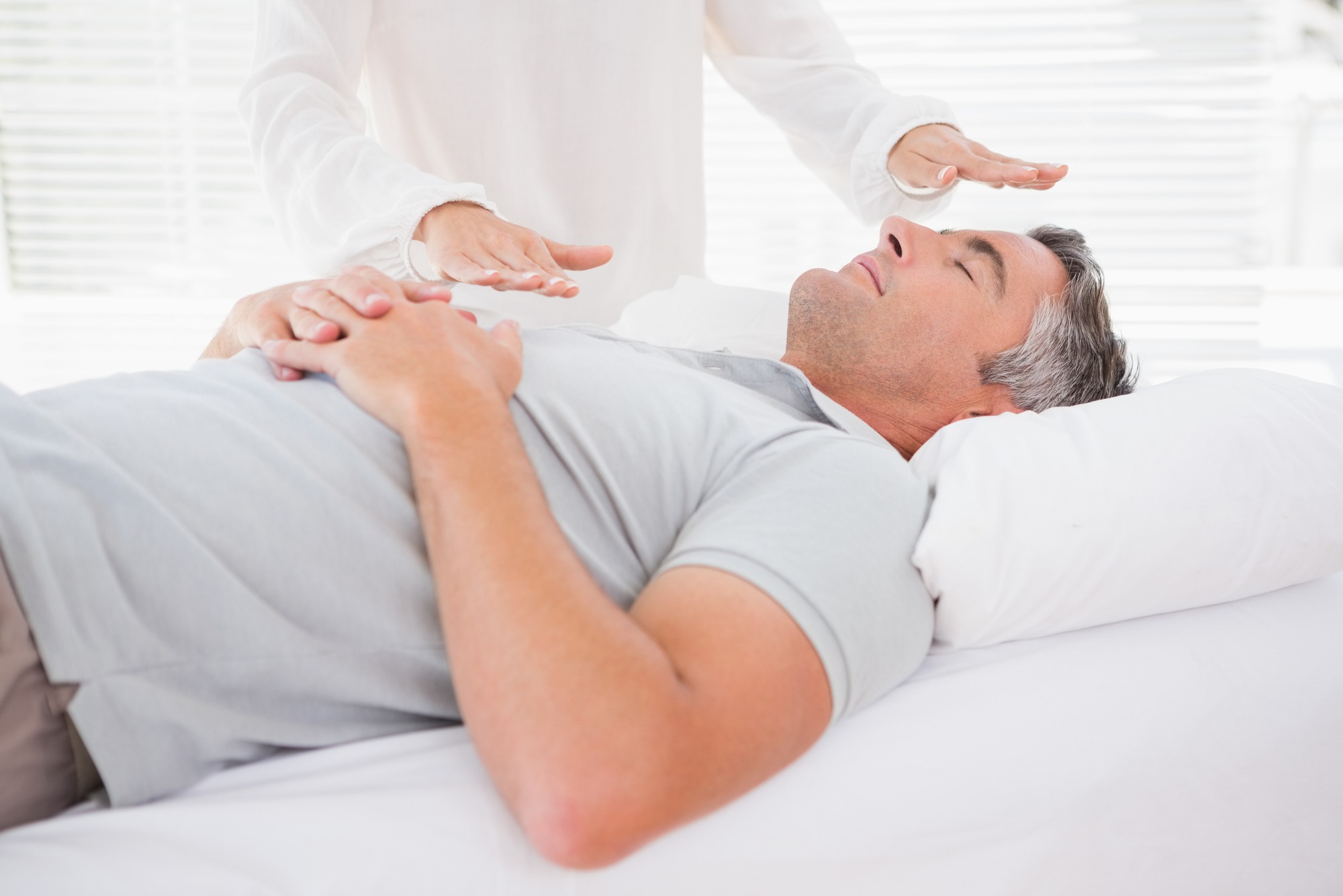 A practitioner is performing energy healing on a relaxed male client lying on a bed. Soft lighting and a peaceful setting enhance the calming atmosphere.