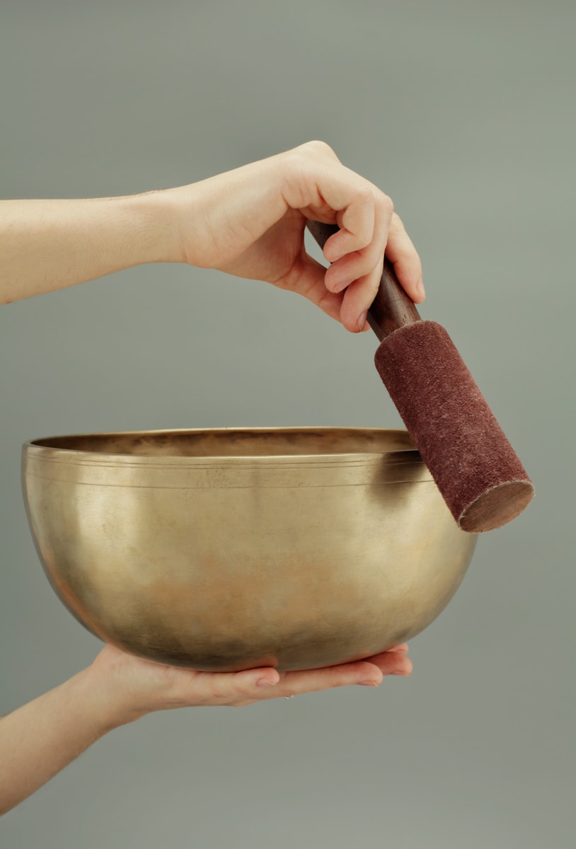A hand holds a striking tool above a shiny brass singing bowl, demonstrating sound therapy practice. The image highlights mindfulness and relaxation techniques.